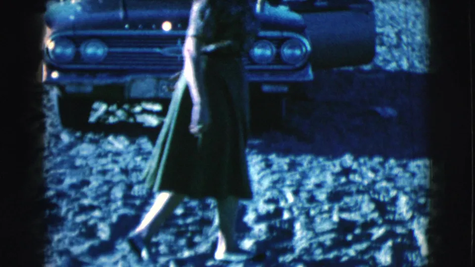 A woman in a green skirt walks across sandy ground in Clarkdale, Arizona, with a parked 1960s car behind her.