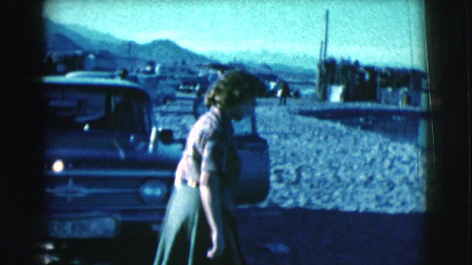 The woman bends slightly while walking beside a parked car, with sandy ground, low shoreline, and mountains visible in Clarkdale.
