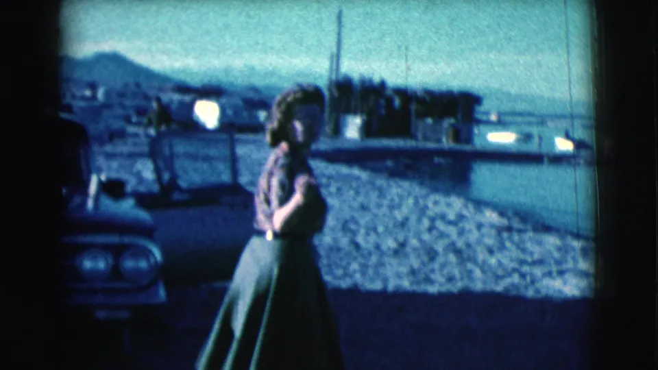 A woman in a green skirt walks near the front of a parked car, with water, sandy ground, and mountains in the 1961 Arizona background.
