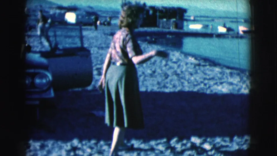 In 1961 Clarkdale, Arizona footage, a woman in a patterned blouse and green skirt gestures beside a car on a pebbled shore.