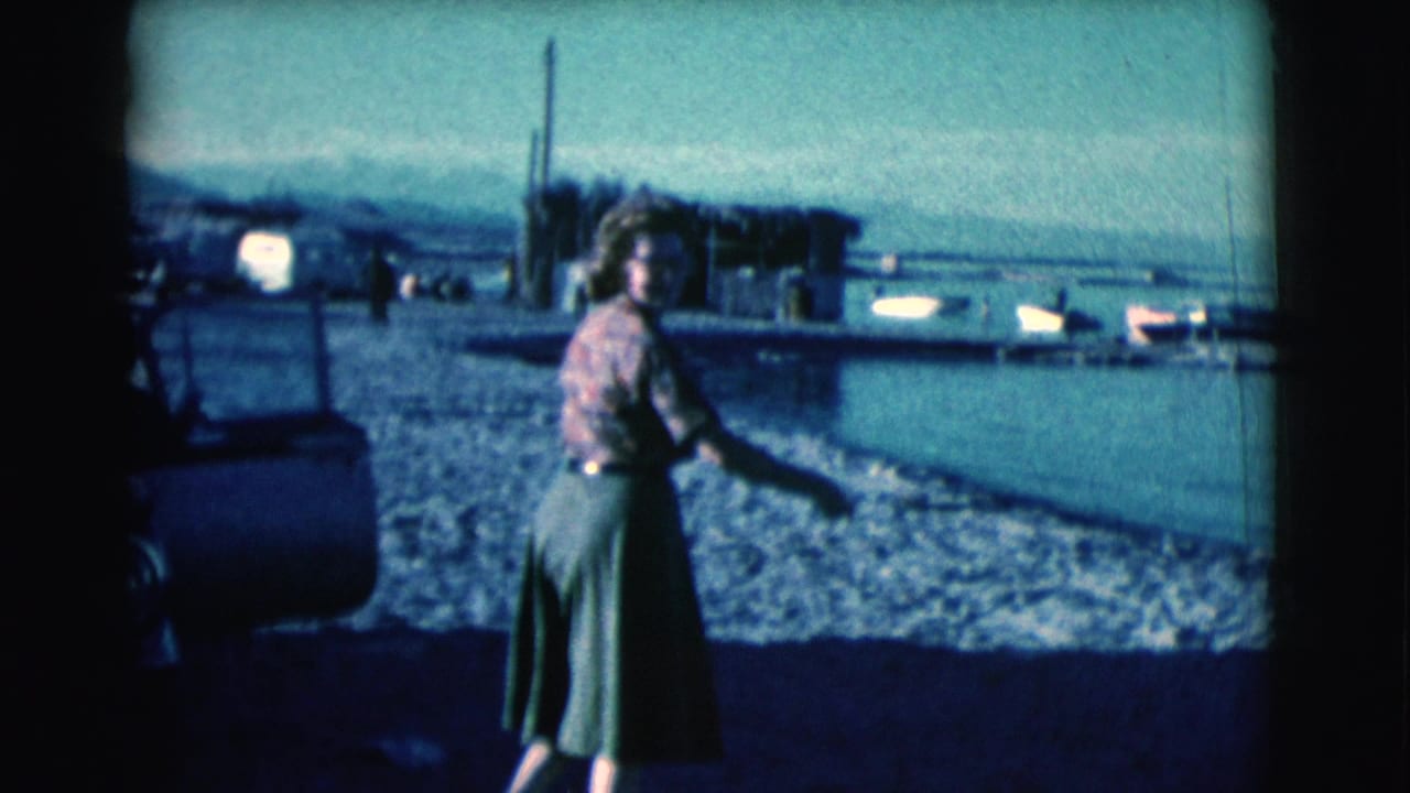Woman walks along the shore with water and low structures in the background.