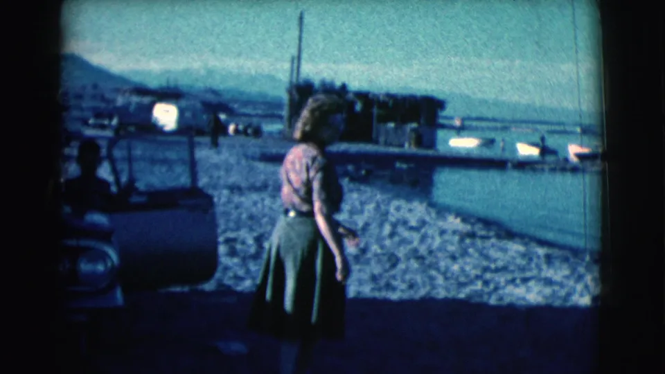 A woman in a green skirt faces the shoreline in 1961 Clarkdale footage, framed with a parked car, water, and distant hills.