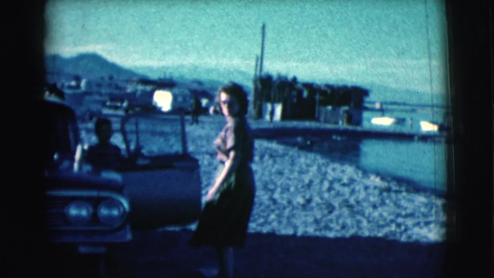 Near the end of the clip, the woman glances back toward the camera beside the car, with water and distant hills behind her.
