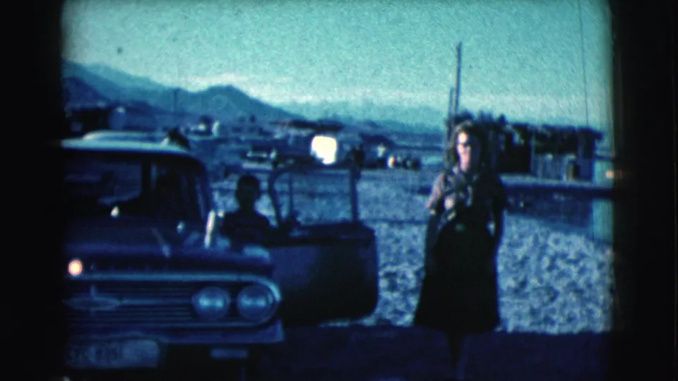 The woman moves along the shoreline beside a 1960s car, with a seated man partly visible through the open door.