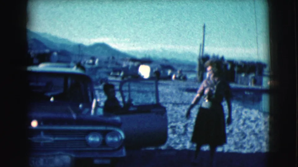 A woman pauses beside the open car door near the Clarkdale waterfront, with distant structures and mountains under hazy light.