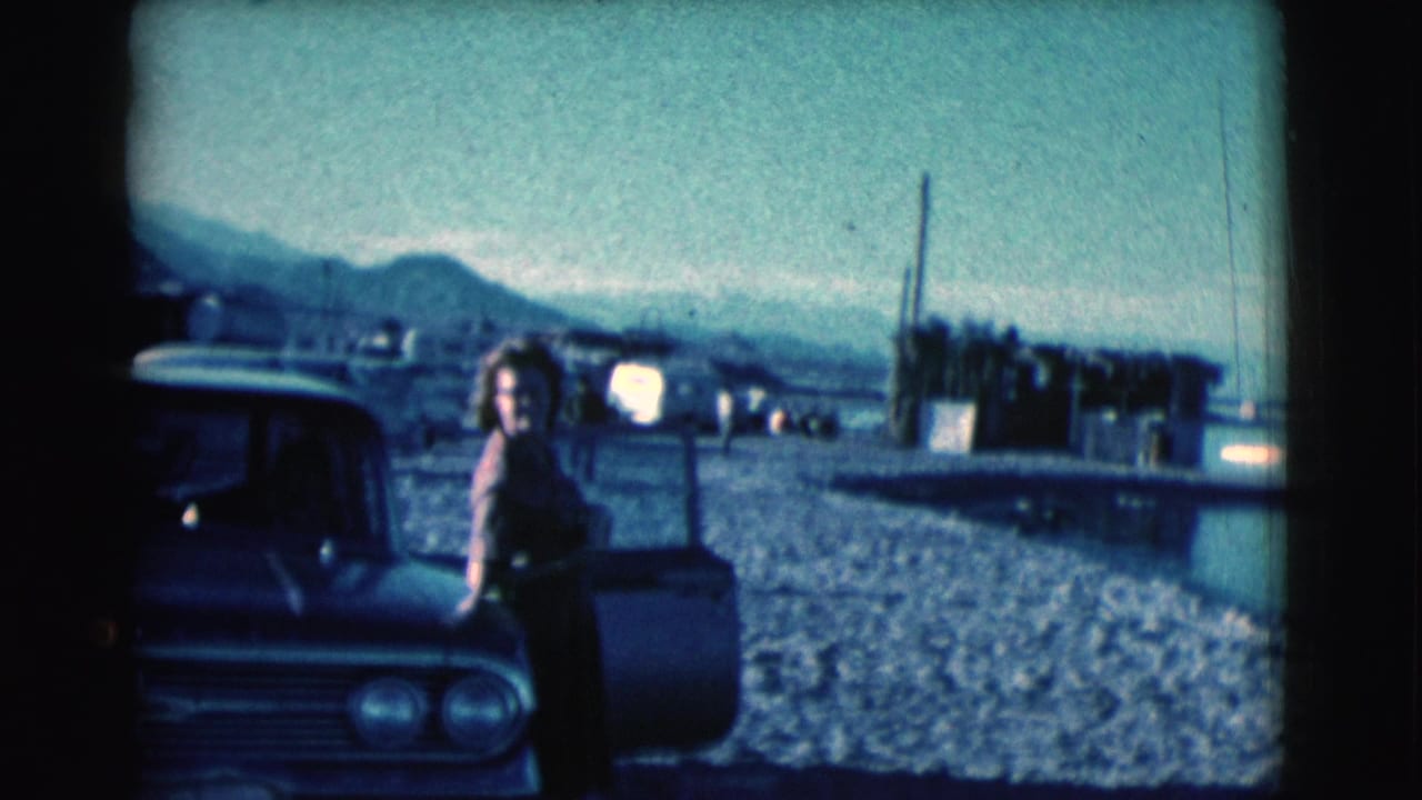 Woman stands beside a dark classic car on a gravel lot near water and mountains.