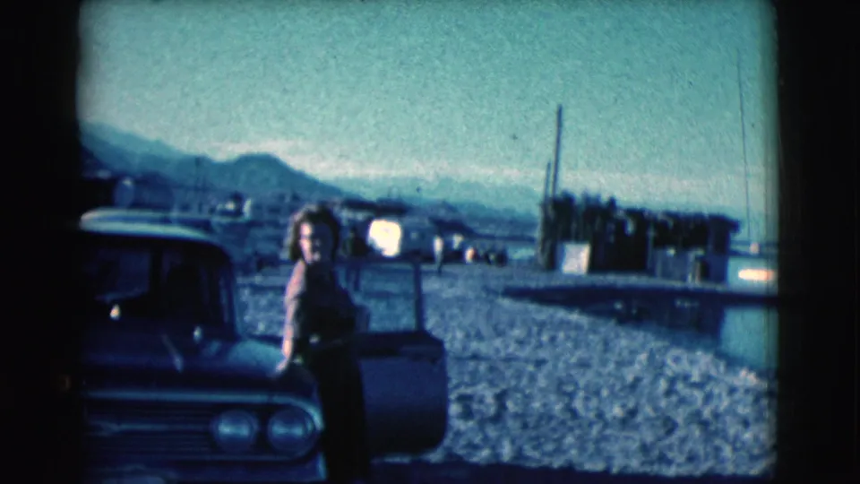 In Clarkdale, Arizona, a woman stands by an open classic car door on a gravel shoreline, with water and mountains behind her.