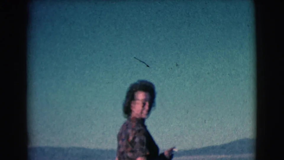 Adult woman in sunglasses and a patterned dress stands near the water in 1961 home-movie footage identified as Clarkdale, Arizona.