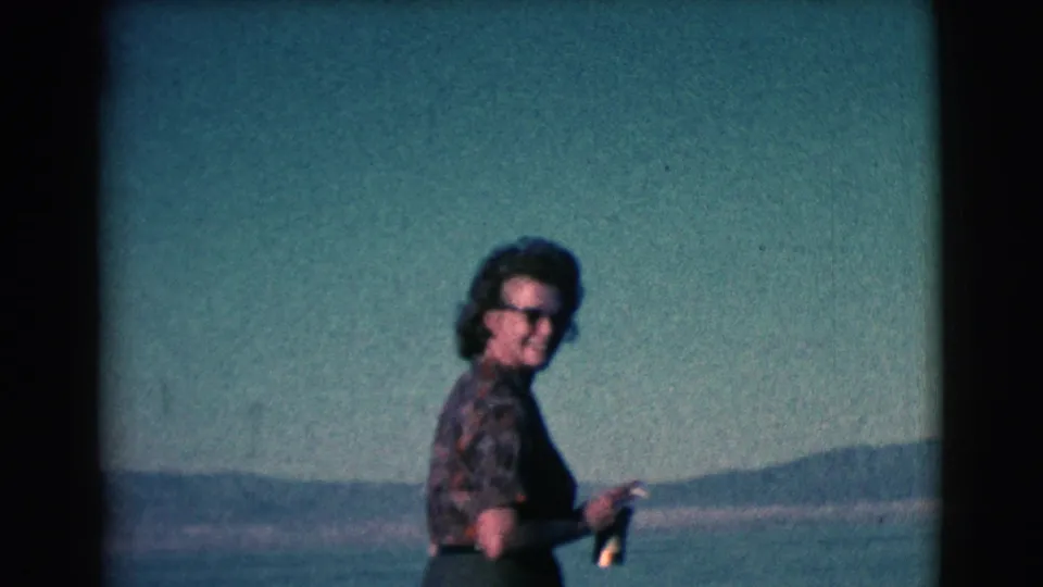 A woman in sunglasses and patterned clothing walks along the shoreline in faded 8mm footage from the 1961 Clarkdale clip.