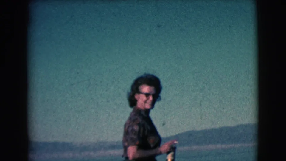 Adult woman in sunglasses is framed against water and distant hills in soft, color-faded 8mm home-movie footage.