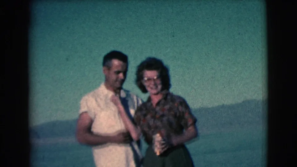 In this 1961 outdoor home-movie frame, the man and woman look down toward the gold-colored cup during the handoff.
