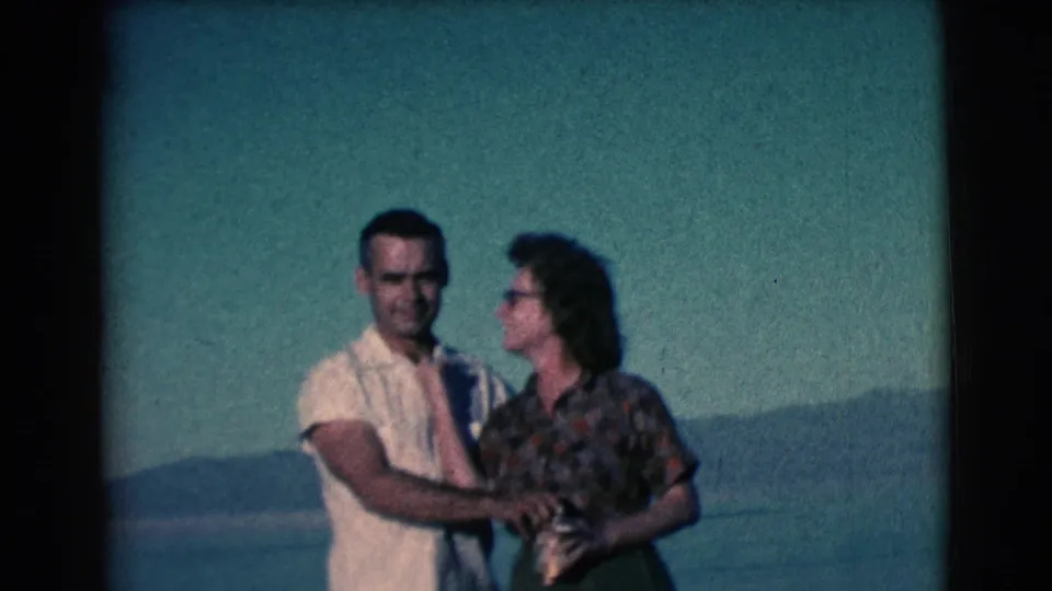 The man reaches for the gold-colored cup as the woman turns toward him, framed against water and hills in the 1961 Clarkdale footage.