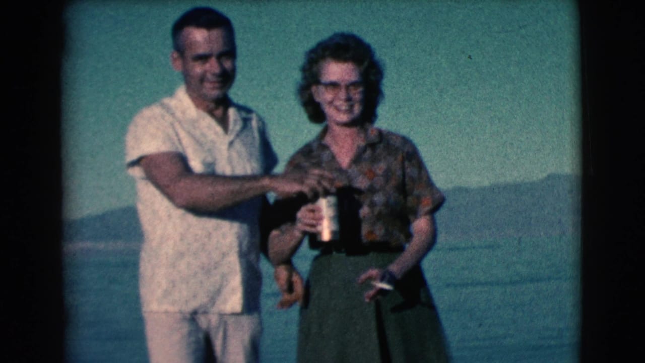 Smiling man and woman stand by water holding drinks with hills in the background.