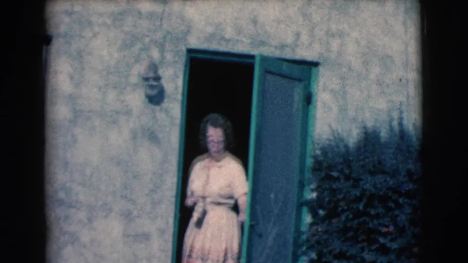 A woman in a pale dress appears at an open green doorway, smiling toward the camera in the 1961 Clarkdale family footage.