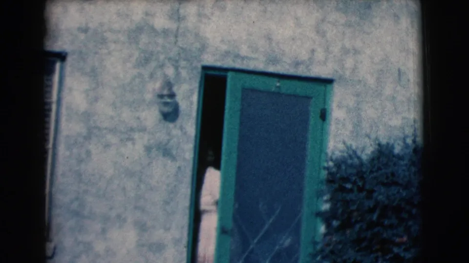 The woman is partly hidden behind the green screen door as the handheld camera holds on the home entrance.
