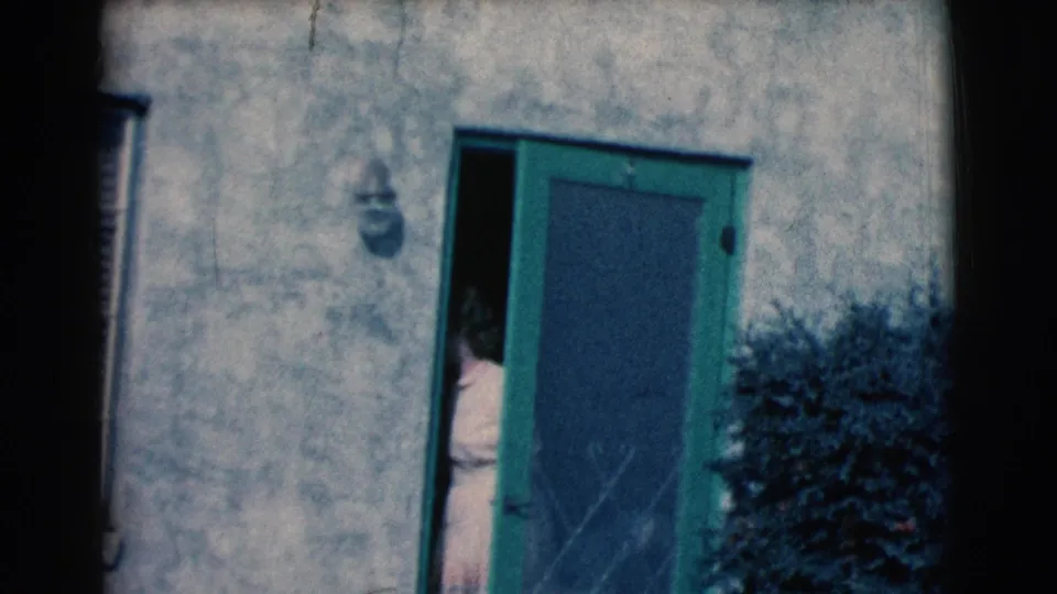 In a 1961 Clarkdale, Arizona home-movie frame, a woman in a light dress is partly visible as she begins to step through a green screen door set in a stucco wall.