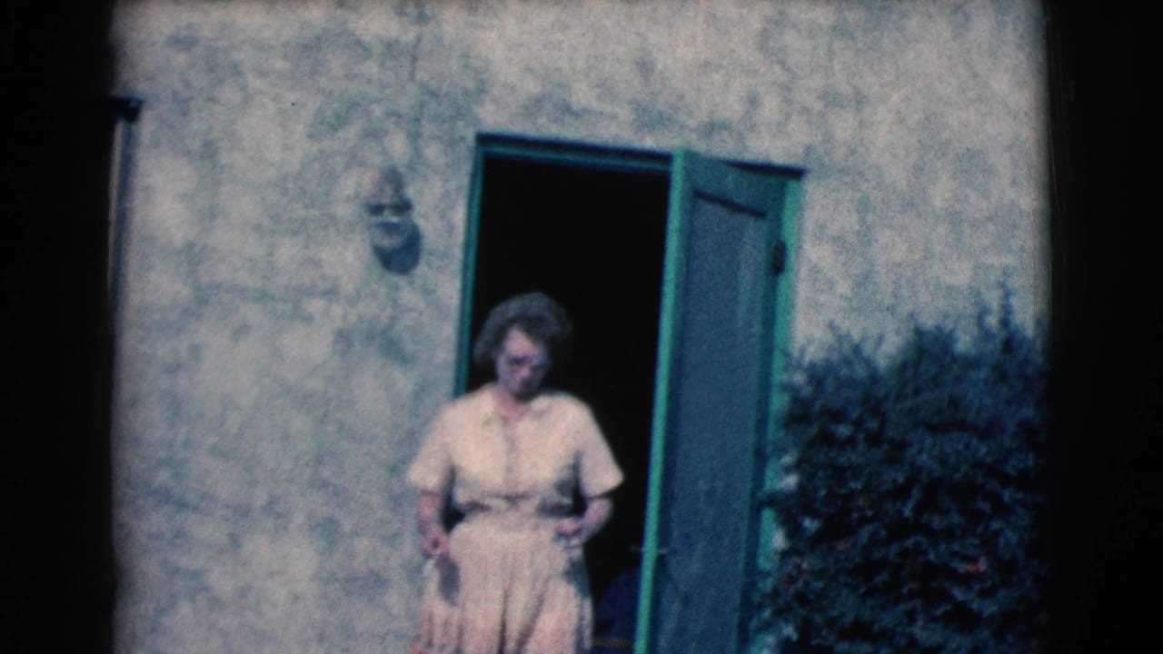 A woman in a light dress and glasses stands just outside an open green screen door on a sunlit stucco house exterior in the supplied 1961 Clarkdale, Arizona footage.