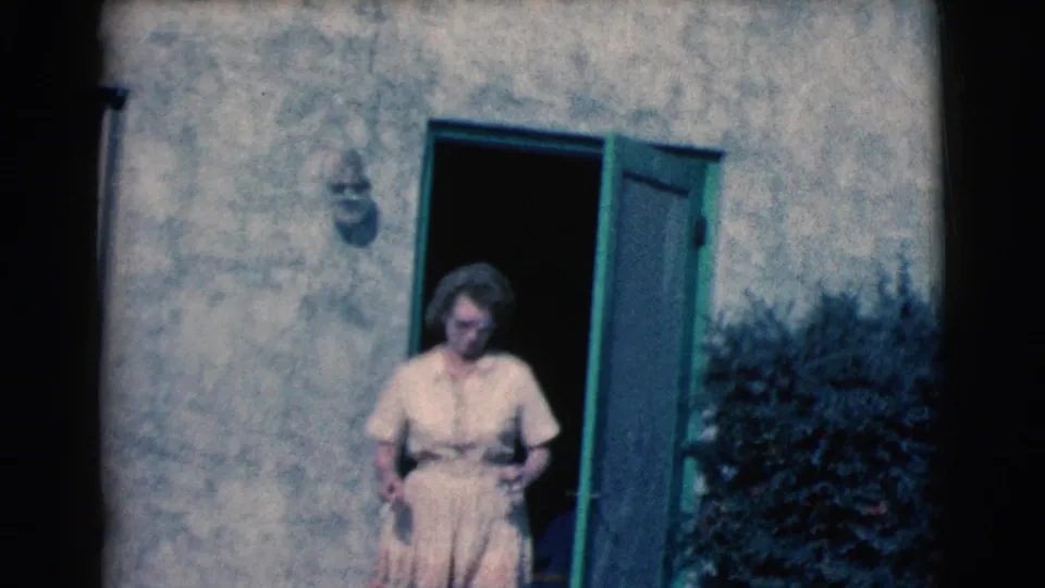 A woman in a light dress and glasses stands just outside an open green screen door on a sunlit stucco house exterior in the supplied 1961 Clarkdale, Arizona footage.