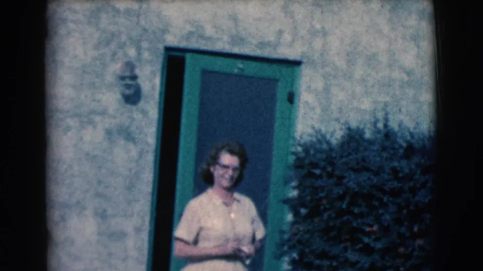 The woman stands near the green screen door, facing the camera with her hands together, with stucco wall texture and a dark shrub visible behind her.