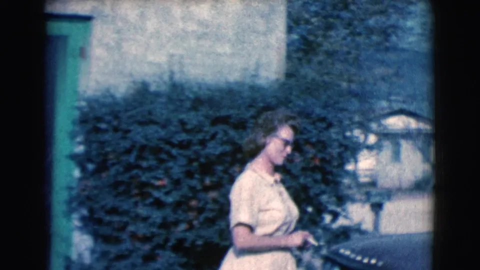 The woman walks away from the green doorway beside dark shrubbery, holding a small object that appears consistent with the cigarette noted in the clip metadata.