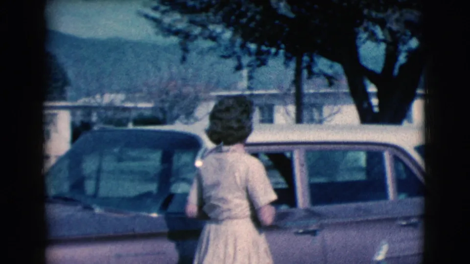 The woman reaches the station wagon in a 1961 Clarkdale residential scene, with low stucco homes, trees, and mountains visible in the background.