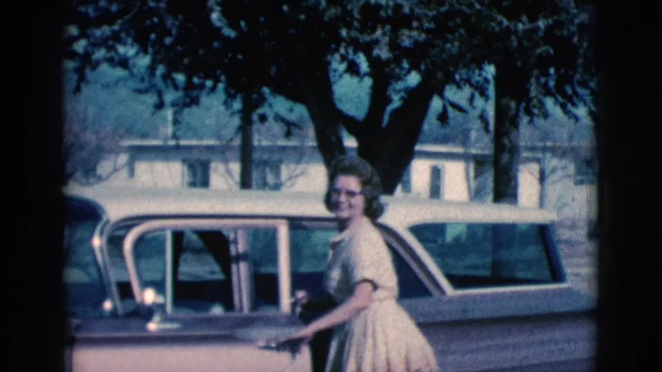 The woman looks back toward the camera next to the station wagon, with her face visible against a backdrop of trees, houses, and mountain slopes.