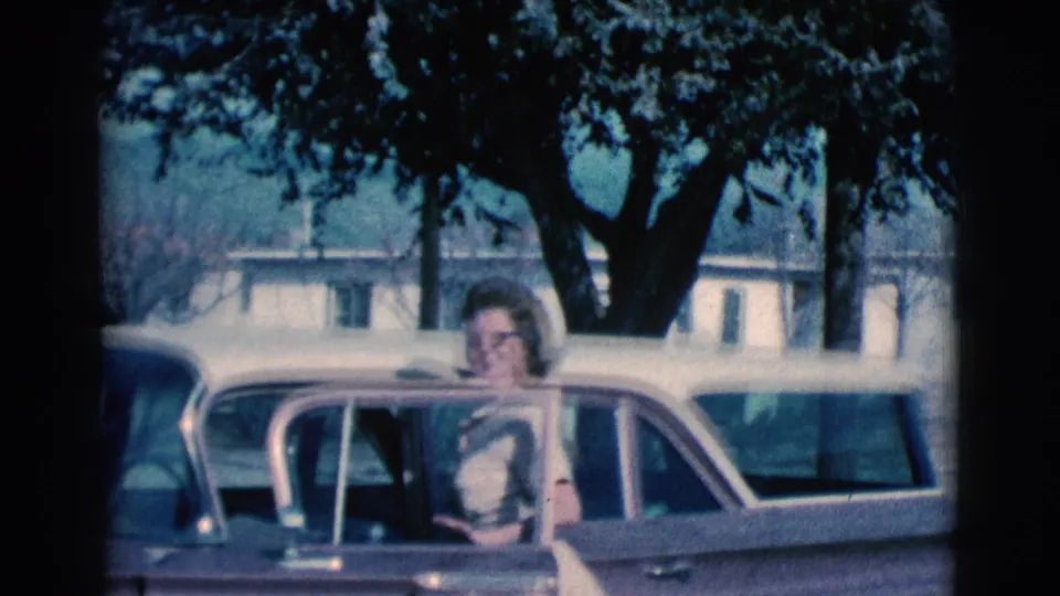 In 1961 Clarkdale, Arizona, a woman in glasses smiles beside an open vintage car door with trees and low buildings behind her.