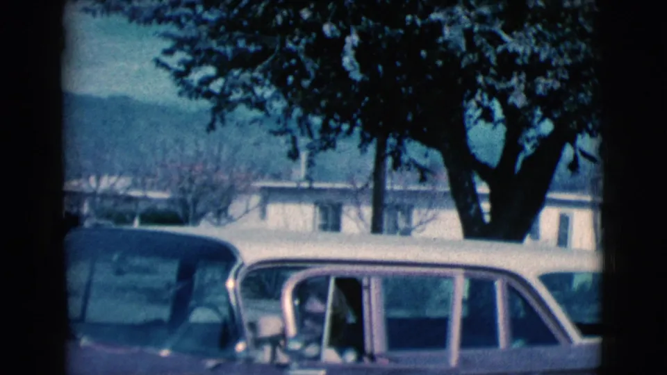 The woman leans into the vintage car in the Clarkdale home-movie scene, with trees, a parked car, and distant hills visible.