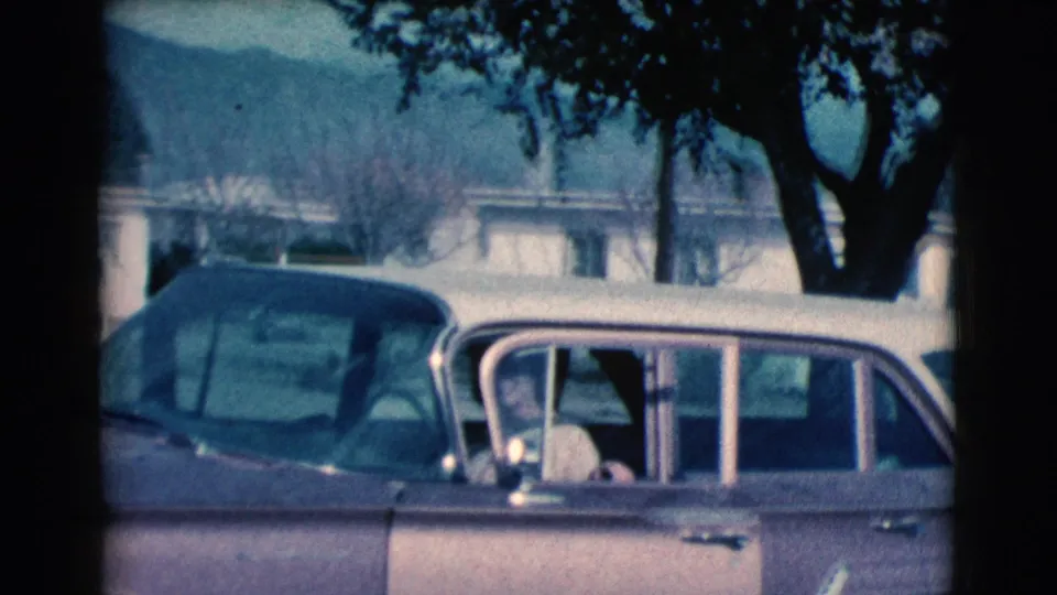 A side view of the vintage car shows the woman mostly inside, with another parked car, trees, and low buildings in the background.