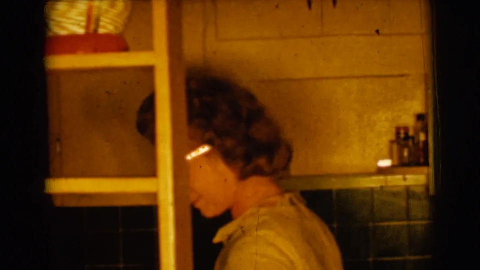 A woman stands in a warmly lit 1961 kitchen, seen in profile with a cigarette and cabinets behind her.