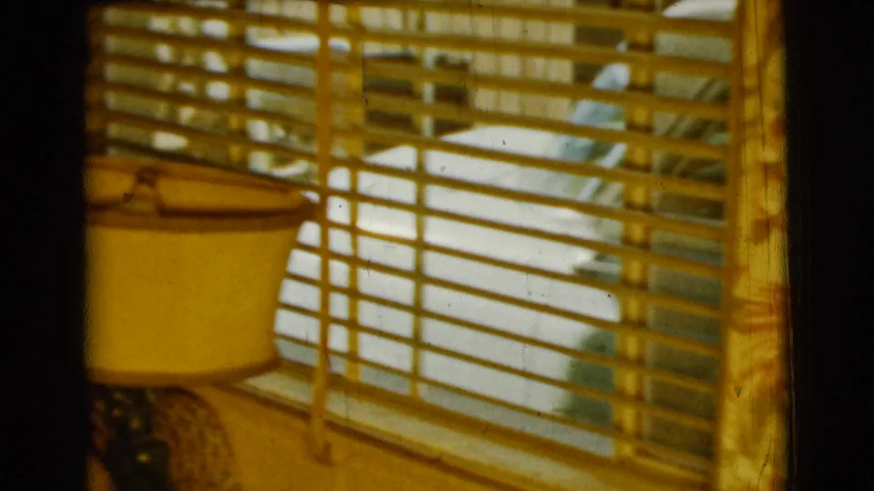 The camera shifts toward a home interior detail with window blinds, a table lamp and furniture in warm 8mm light.