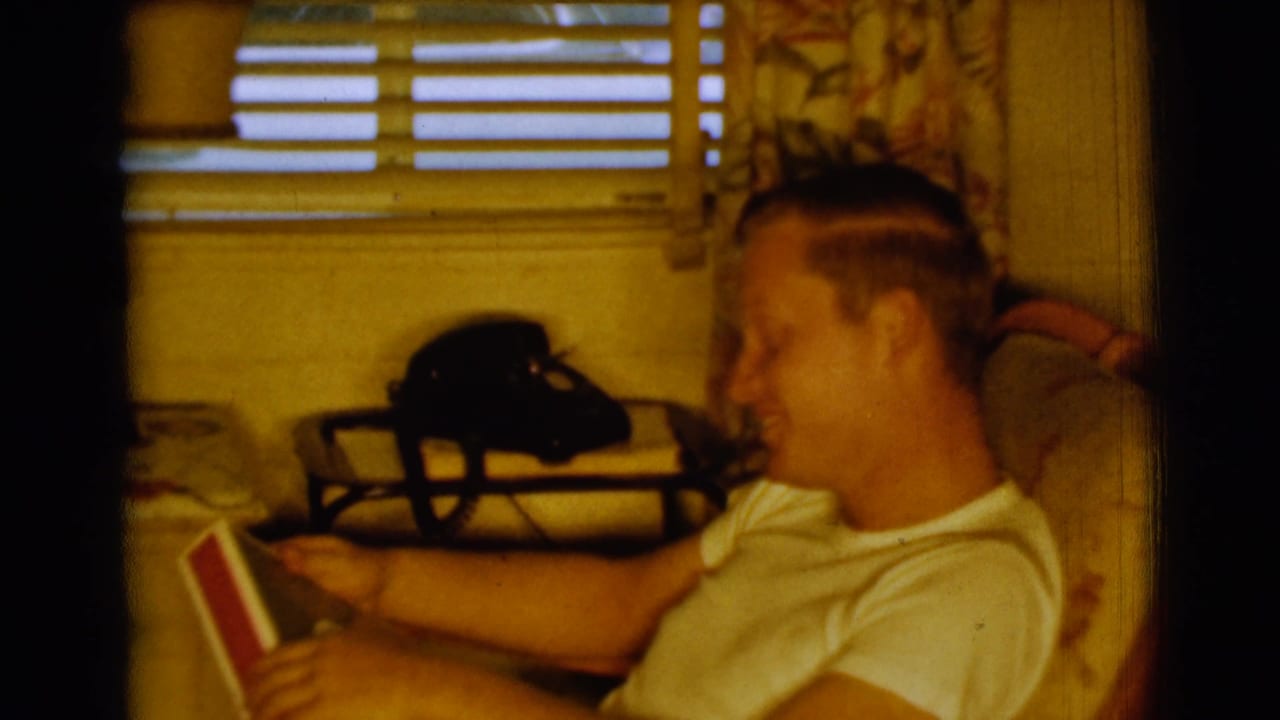 Young man seated indoors holding a record album near window blinds and a rotary telephone.