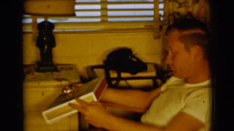 A young man sits in a chair in the Clarkdale 1961 home-movie scene, holding a record album near a lamp, blinds, and rotary telephone.