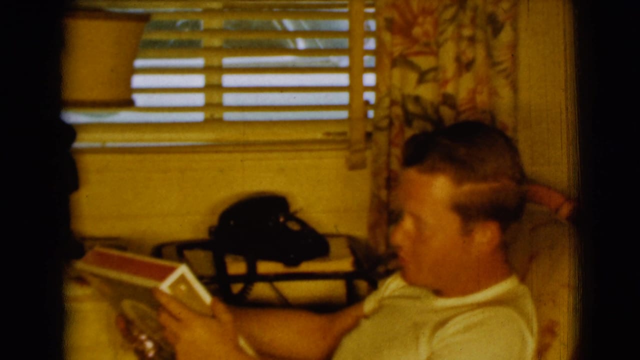 Man in a white T-shirt reads a red-and-white book beside a black rotary phone and window blinds.