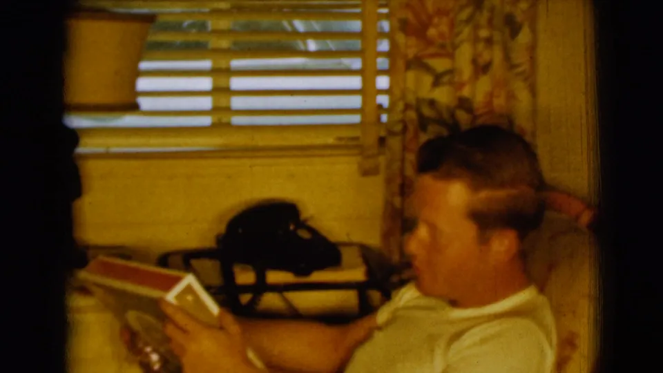 In a 1961 Clarkdale, Arizona home interior, a seated man reads a red-and-white book near a black rotary phone and sunlit blinds.