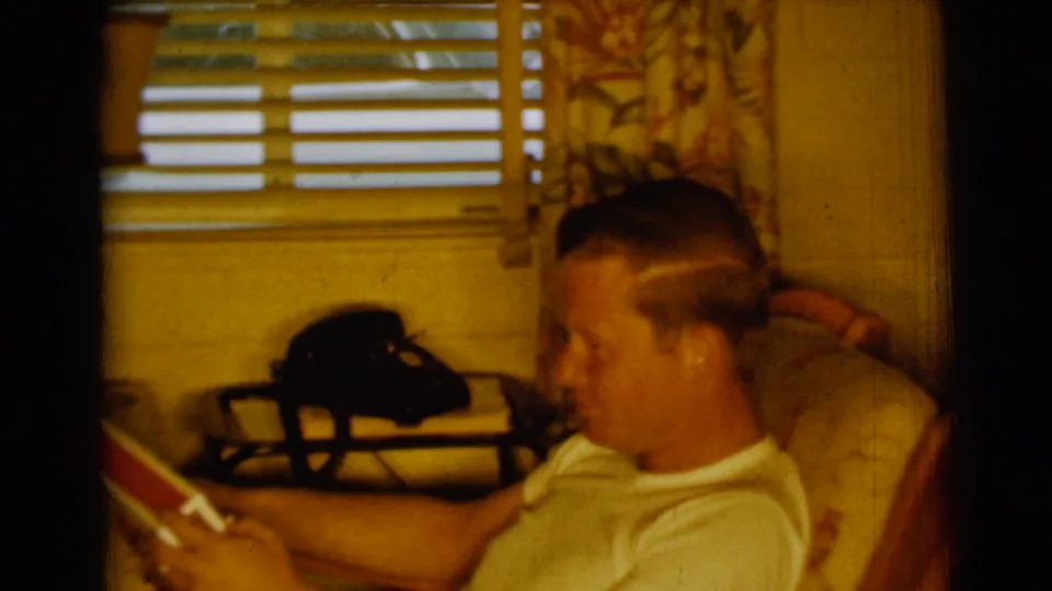 The seated man continues reading in the Clarkdale home, with a black rotary phone, blinds, and patterned curtains visible behind him.