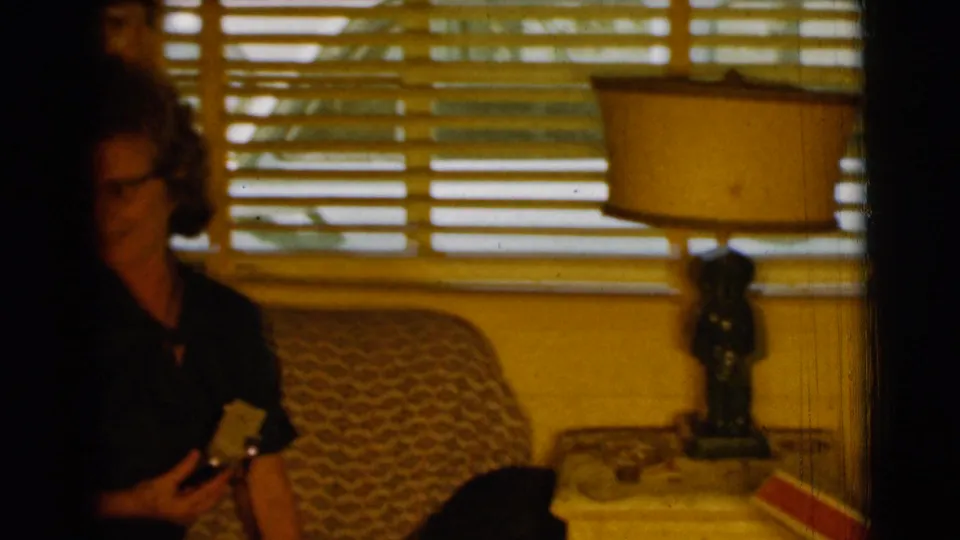 A woman in a dark dress appears in the 1961 Clarkdale interior, holding a small object near a lamp, blinds, and reading material.