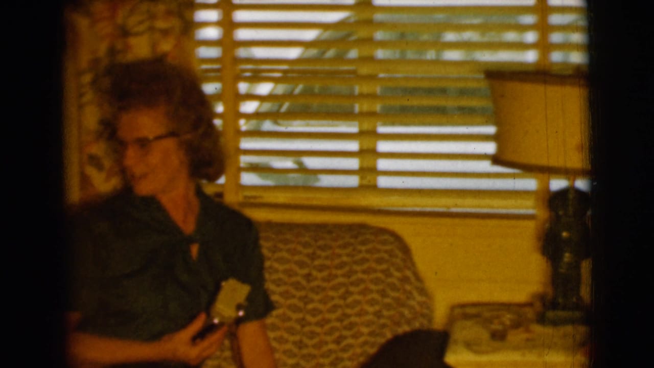 Older woman with glasses sits on a patterned couch holding a small shiny item.