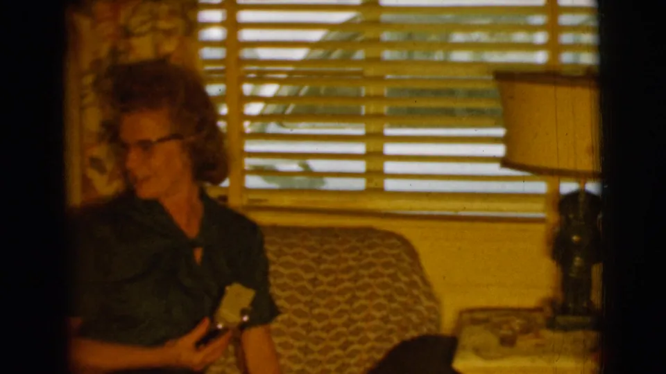 In a Clarkdale, Arizona 1961 home-movie frame, an older woman in glasses sits on a couch near blinds while holding a reflective object.