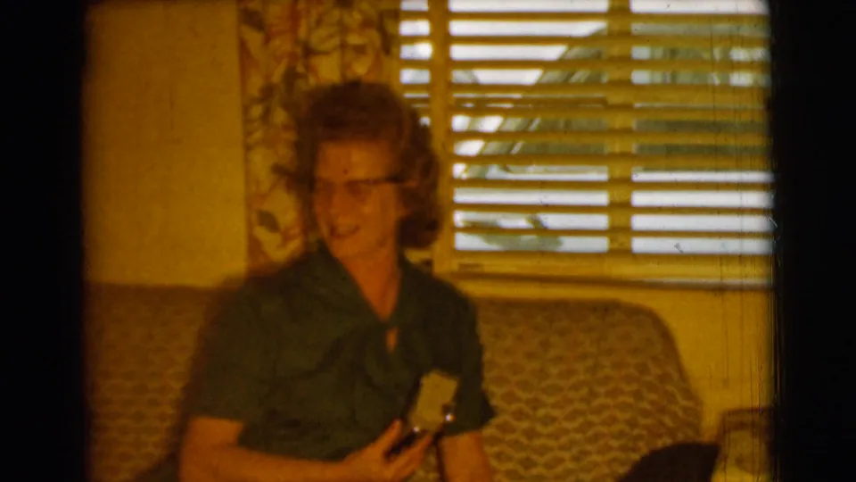 The woman appears to smile or speak while seated in a living room, with blinds and a lamp visible behind her in the 1961 Clarkdale footage.