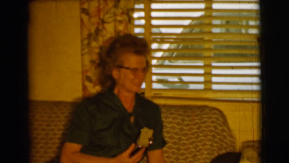 An older woman in glasses sits against a patterned couch in warm 8mm color, looking down toward the object she is holding.