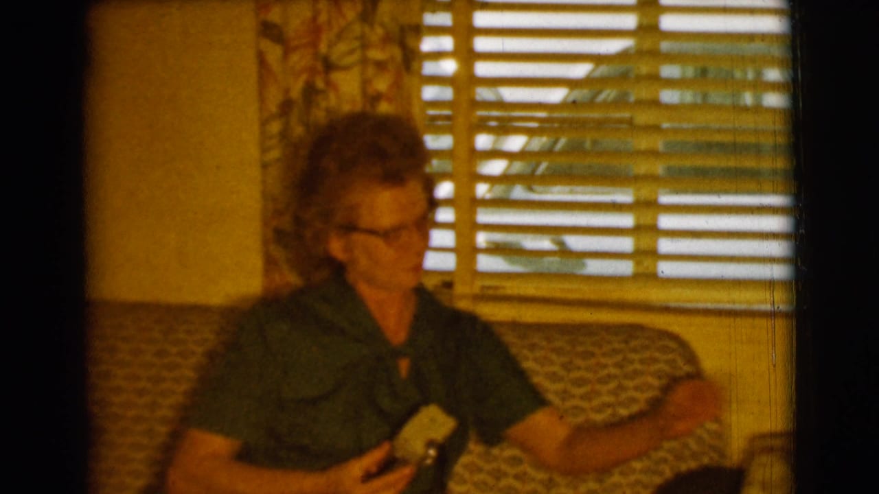 Woman in glasses sits on a patterned sofa holding a cigarette near window blinds.