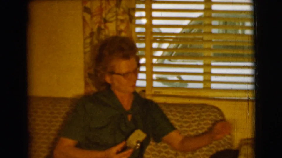 In a 1961 Clarkdale, Arizona home interior, a woman in glasses sits on a patterned sofa holding a cigarette by window blinds.