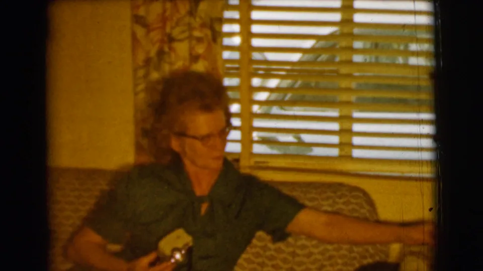 The woman sits by the blinds in the Clarkdale home interior, extending one arm while holding a cigarette.