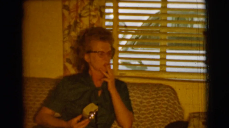 In the 1961 home-movie frame, the woman raises a cigarette toward her mouth while seated on the patterned sofa.
