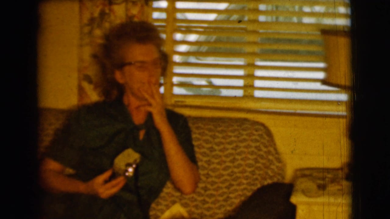 Woman in glasses sits on a patterned sofa smoking and holding a small object.