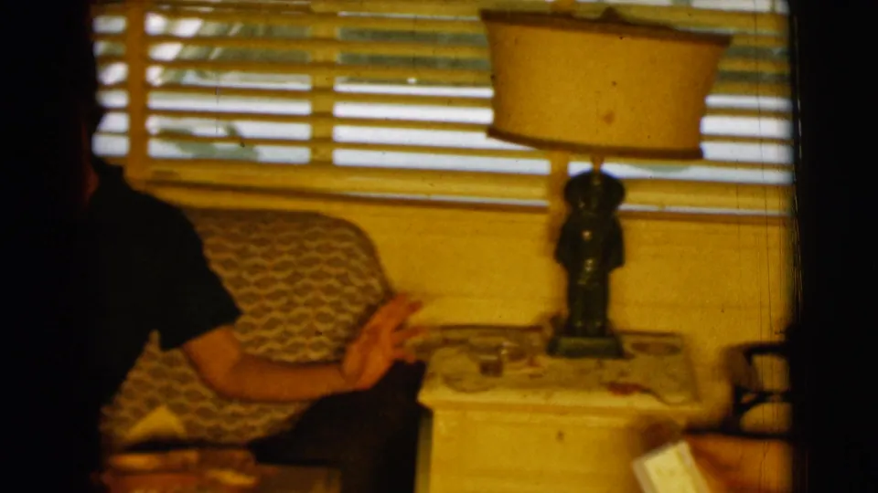 A 1961 Clarkdale, Arizona living-room frame shows a partly obscured seated man near a sofa, side lamp, and slatted blinds.