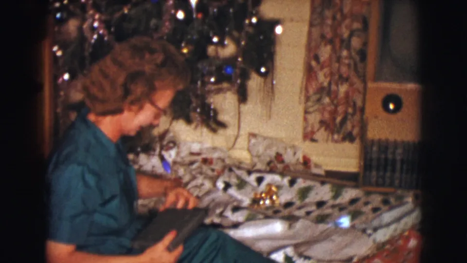 The woman leans over a dark box or package, with wrapping paper and Christmas tree decorations visible around her.