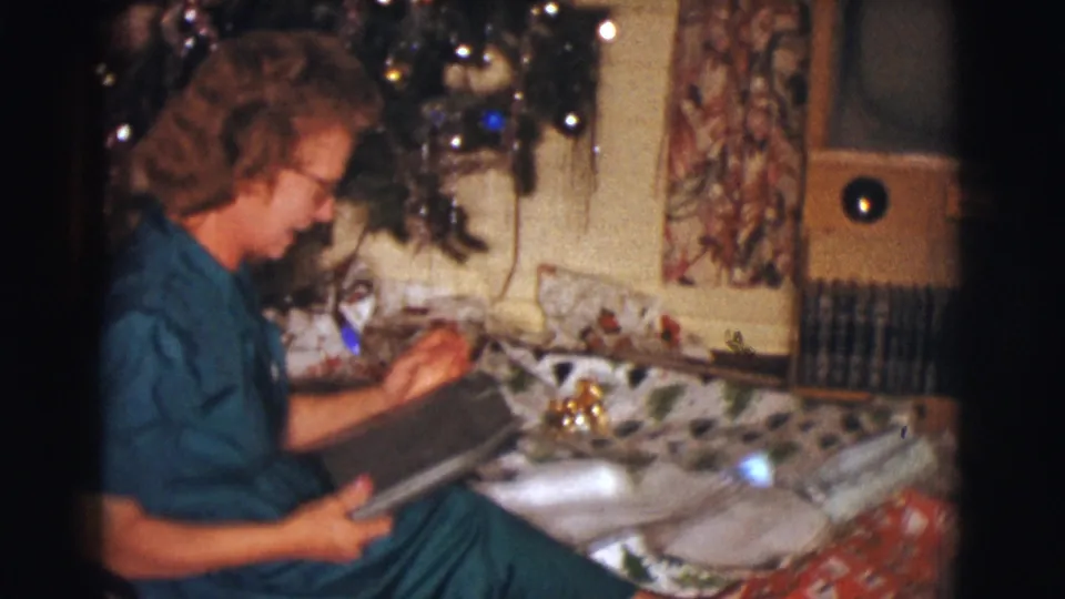 The woman gestures while holding a package beside the decorated Christmas tree, with wrapped gifts and curtains behind her.