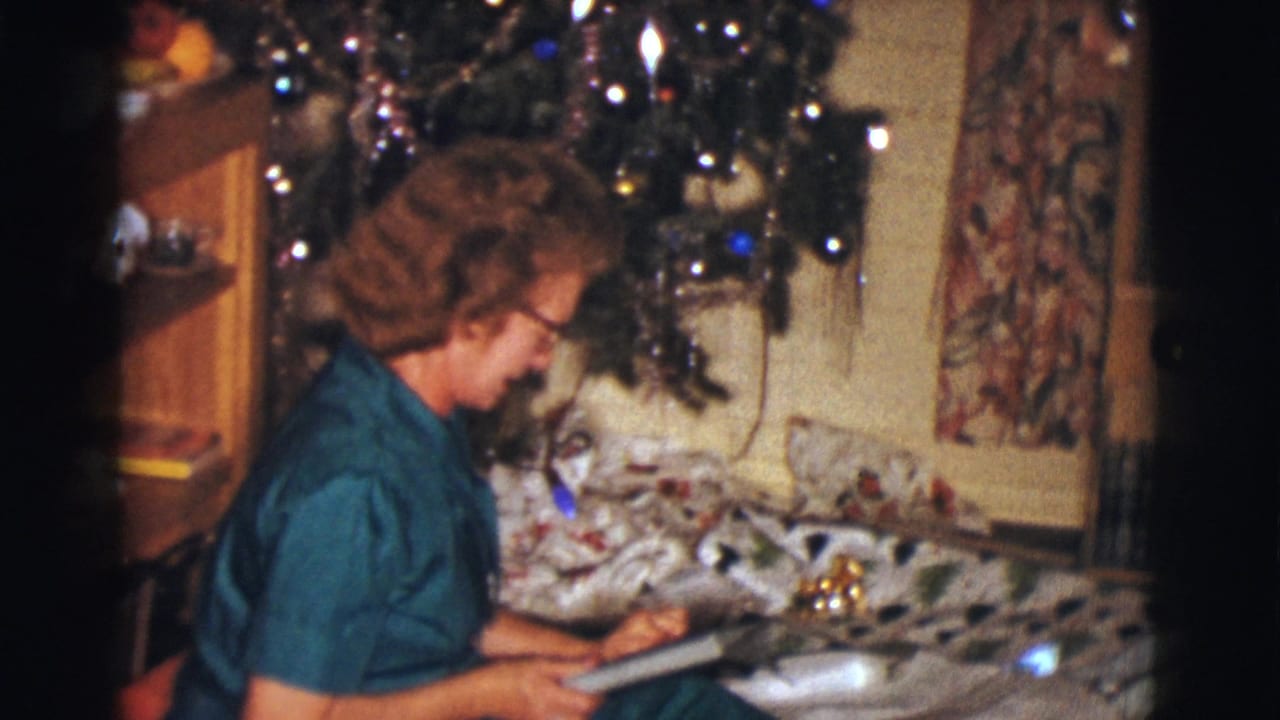 The woman sits in profile near the Christmas tree, focused on wrapping paper or a present in the living room.